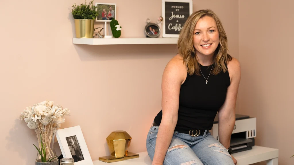 Business Strategist Jennifer Sakowski sits on a desk in her office wearing jeans, a black top and smiling.