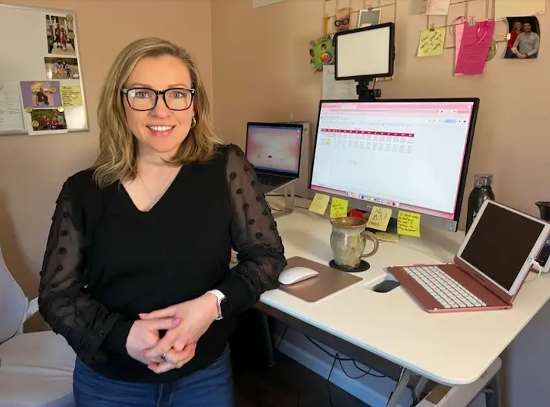 Jennifer Sakowski stands smiling with her hands folded in front of her desk and devices as she works in her office